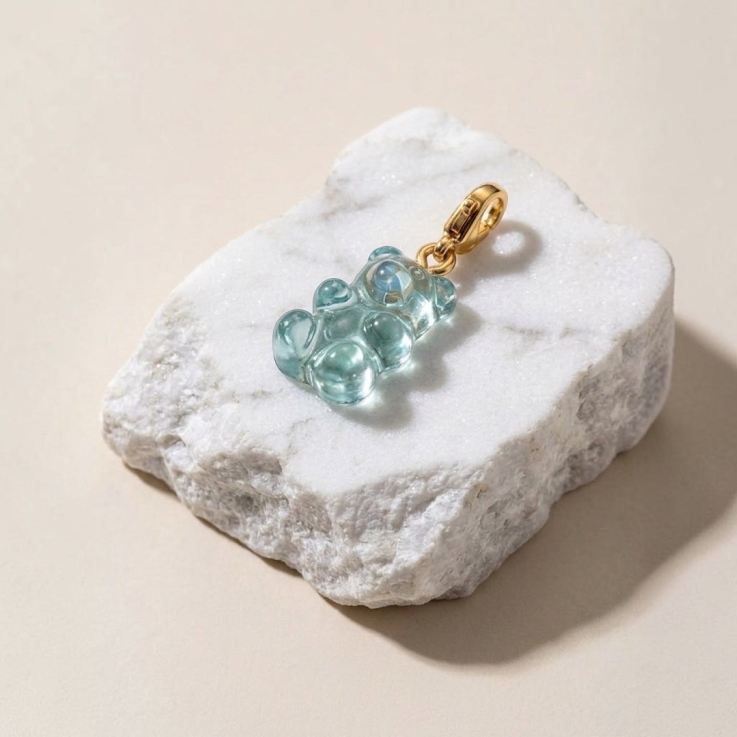Chévereh translucent light blue resin gummy bear charm with a 14K gold-plated lobster clasp resting on a white stone.
