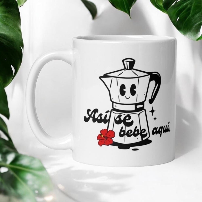 Chévereh Ceramic Mug 11oz with a smiling greca coffee maker, red hibiscus flower, and text "Así se bebe aquí".