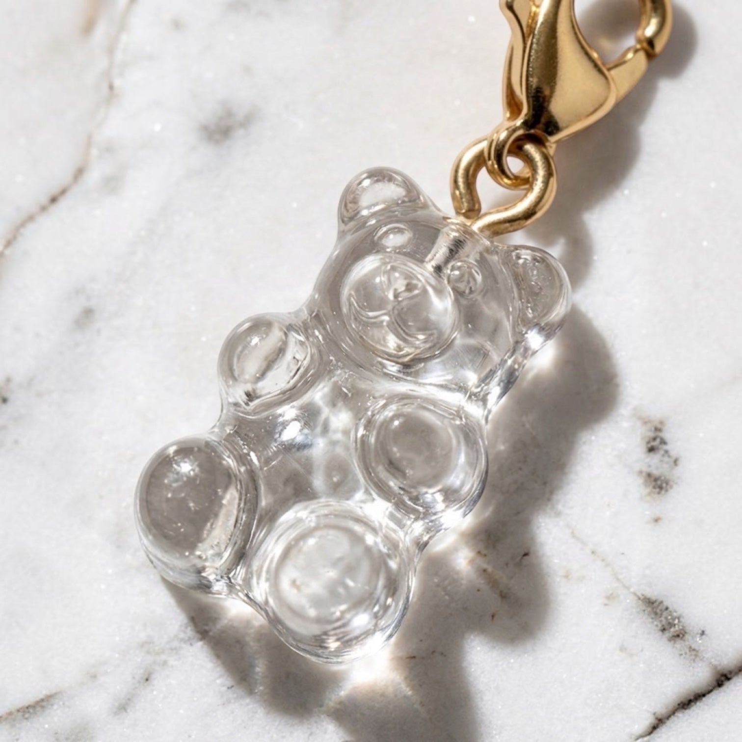 Chévereh clear resin gummy bear charm with a polished 14K gold-plated lobster clasp on a white marble surface.