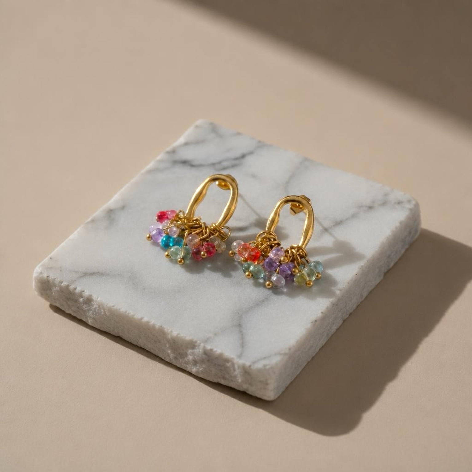 Chévereh gold-tone oval hoop earrings featuring a cluster of vibrant multi-colored beads on a marble display block.