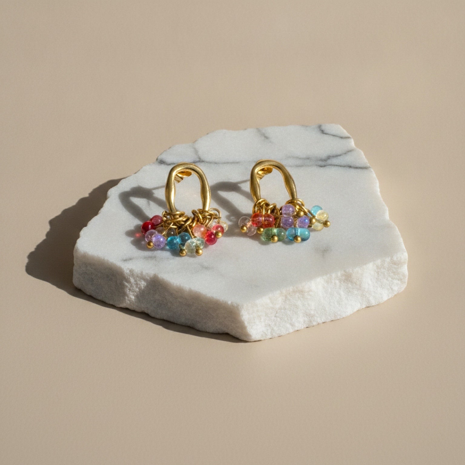 Chévereh gold-tone oval hoop earrings featuring a cluster of vibrant multi-colored beads on a white marble display.