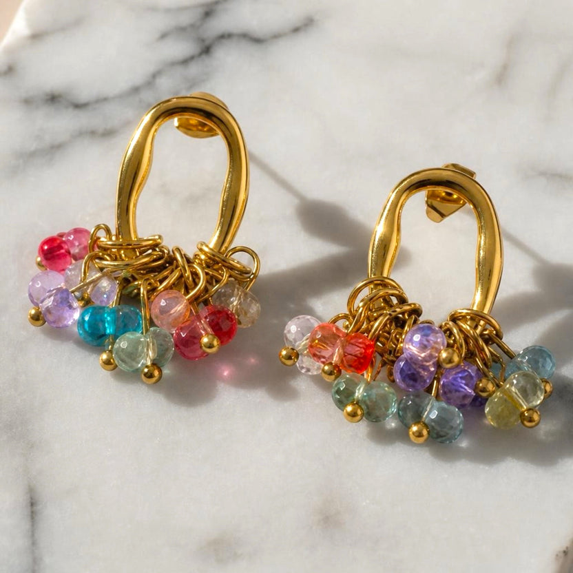 Chévereh gold-tone oval hoop earrings featuring a cluster of vibrant multi-colored faceted beads on a marble surface.