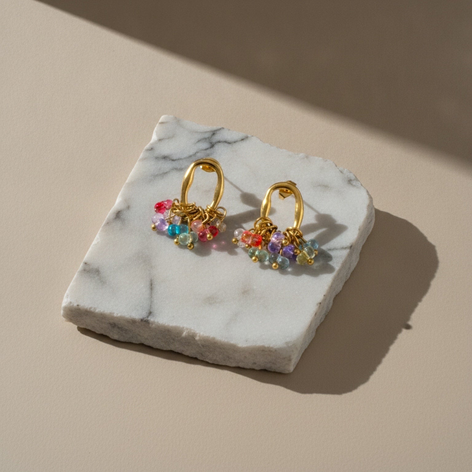 Chévereh gold-tone oval hoop earrings featuring a cluster of vibrant multi-colored beads on a marble display stand.