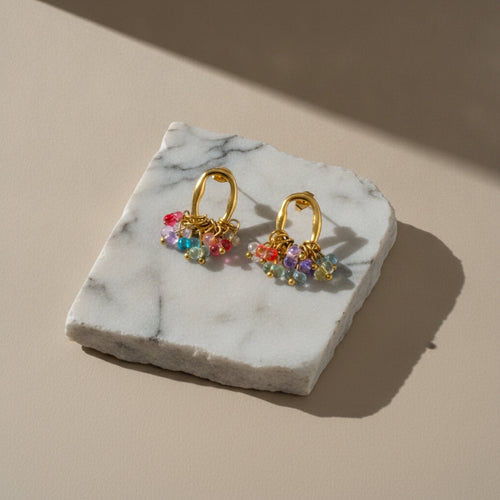 Chévereh gold-tone oval hoop earrings featuring a cluster of vibrant multi-colored beads on a marble display stand.