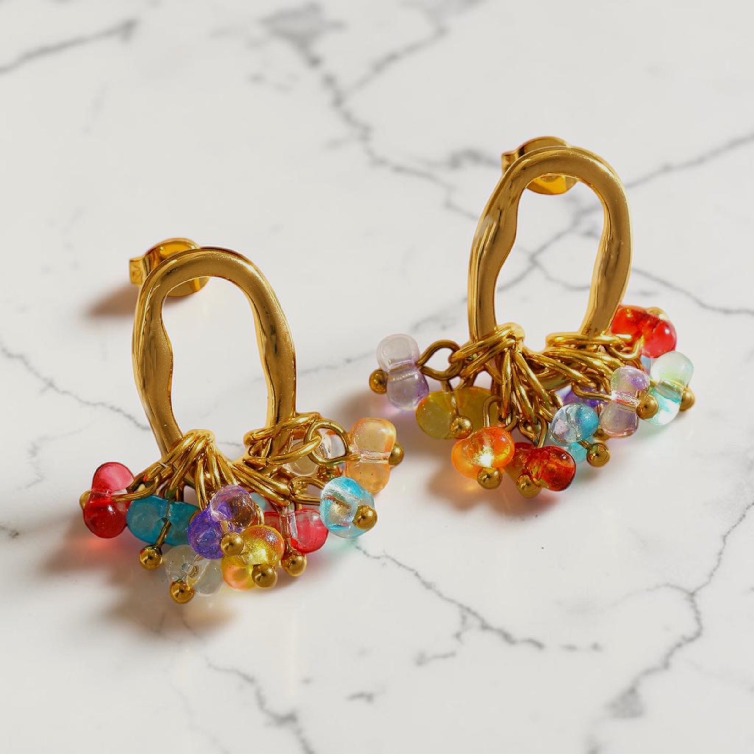 Chévereh gold-tone oval hoop earrings featuring a vibrant cluster of multi-colored glass beads on a marble surface.