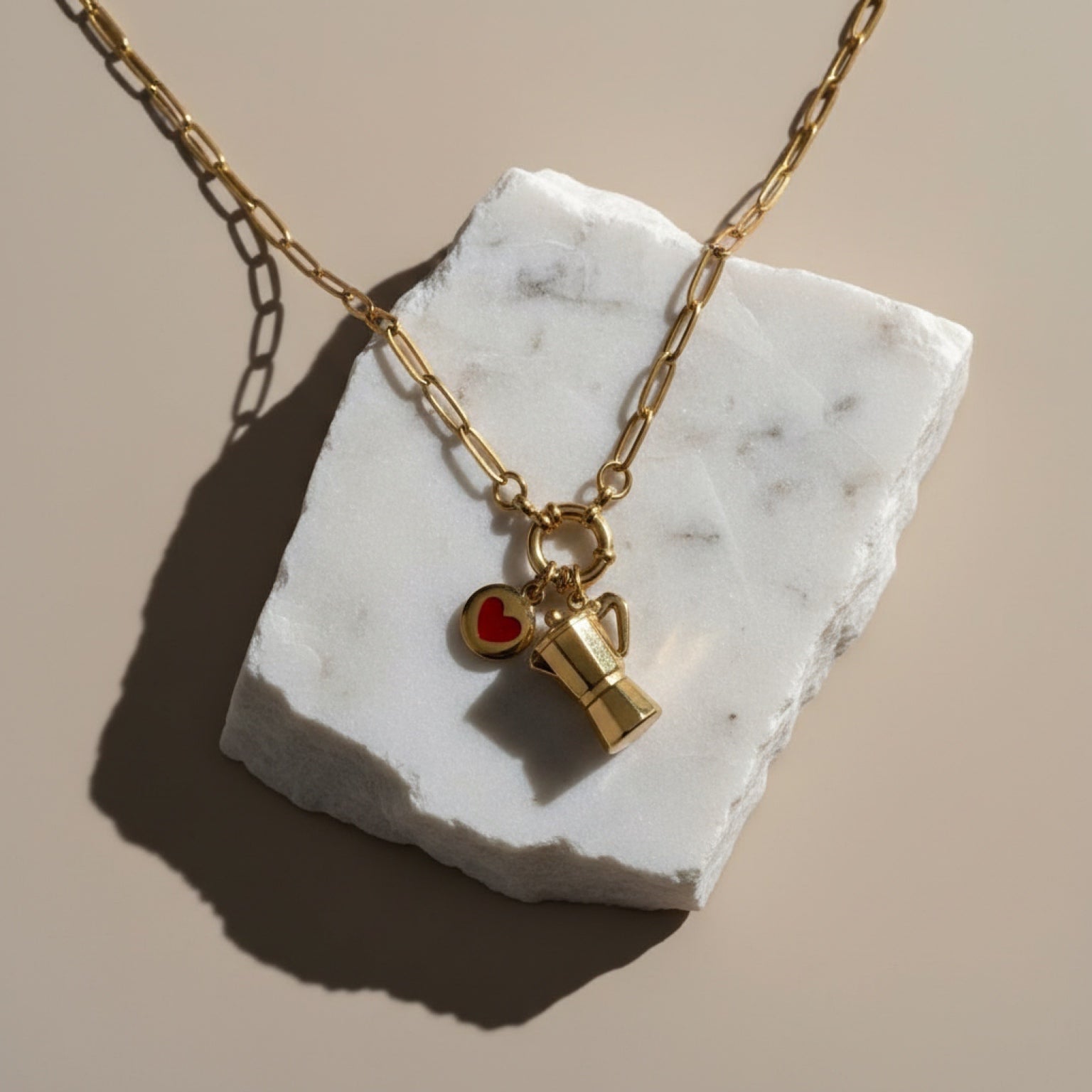 Chévereh gold paperclip chain necklace with moka pot and heart charms displayed on a white marble stone.