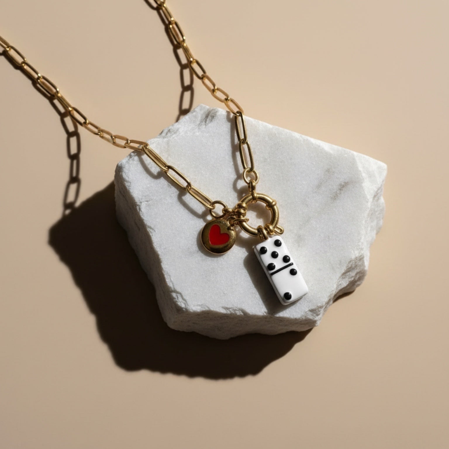 Chévereh gold paperclip chain necklace with domino and heart charms on a white stone display.