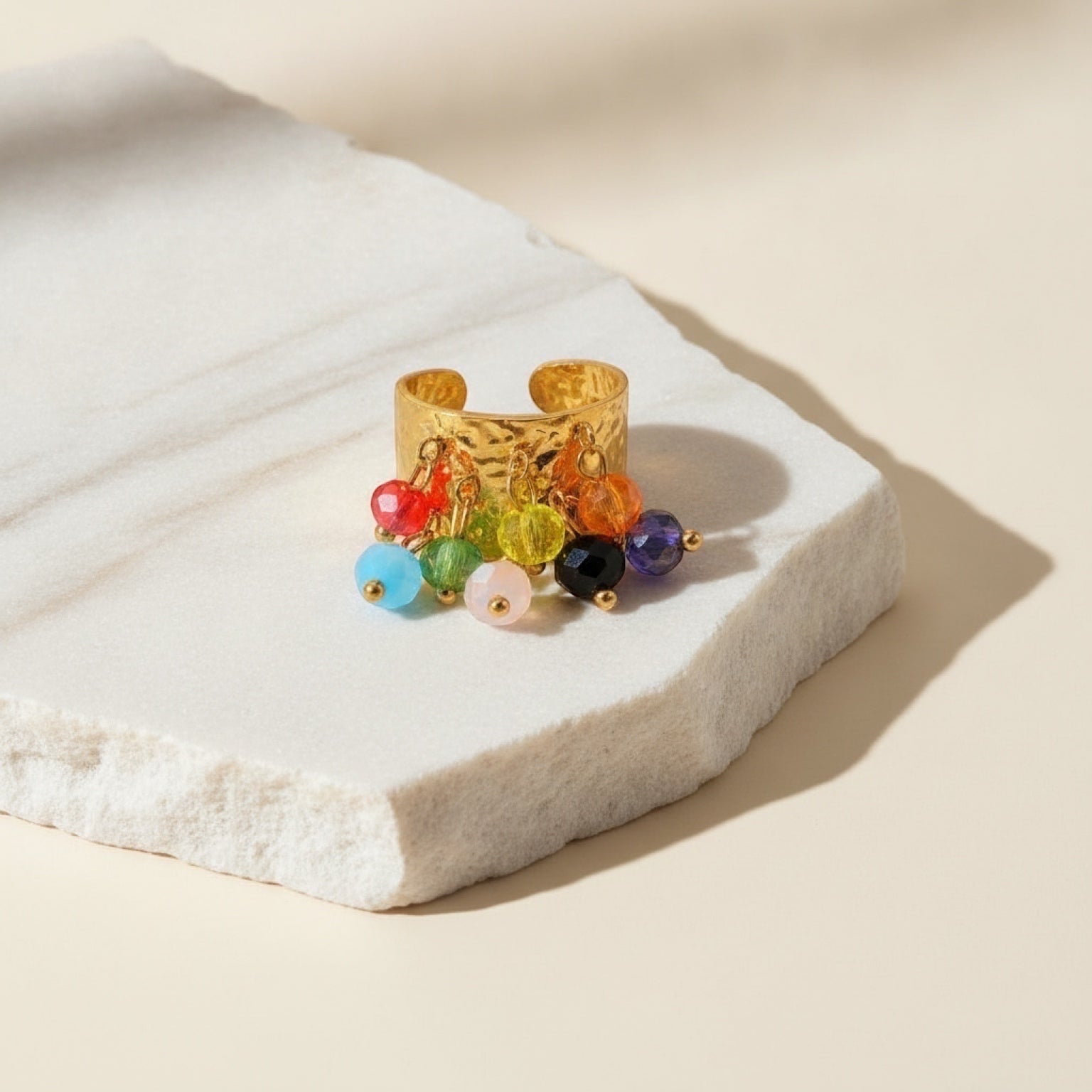 Chévereh gold-toned hammered metal statement ring with dangling multi-colored beaded charms on a stone surface
