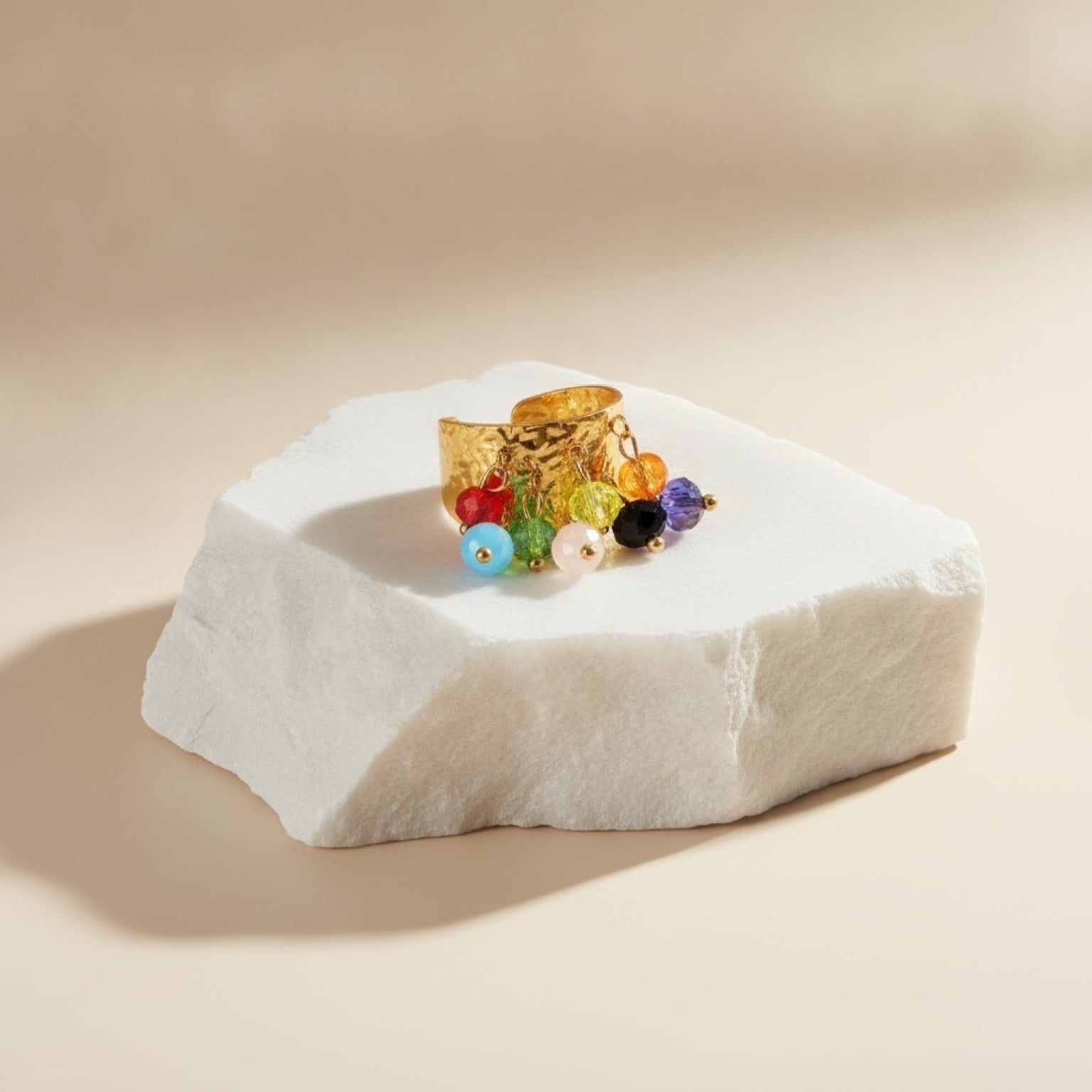 Chévereh gold hammered metal statement ring with dangling multi-colored beaded charms on a white stone display.