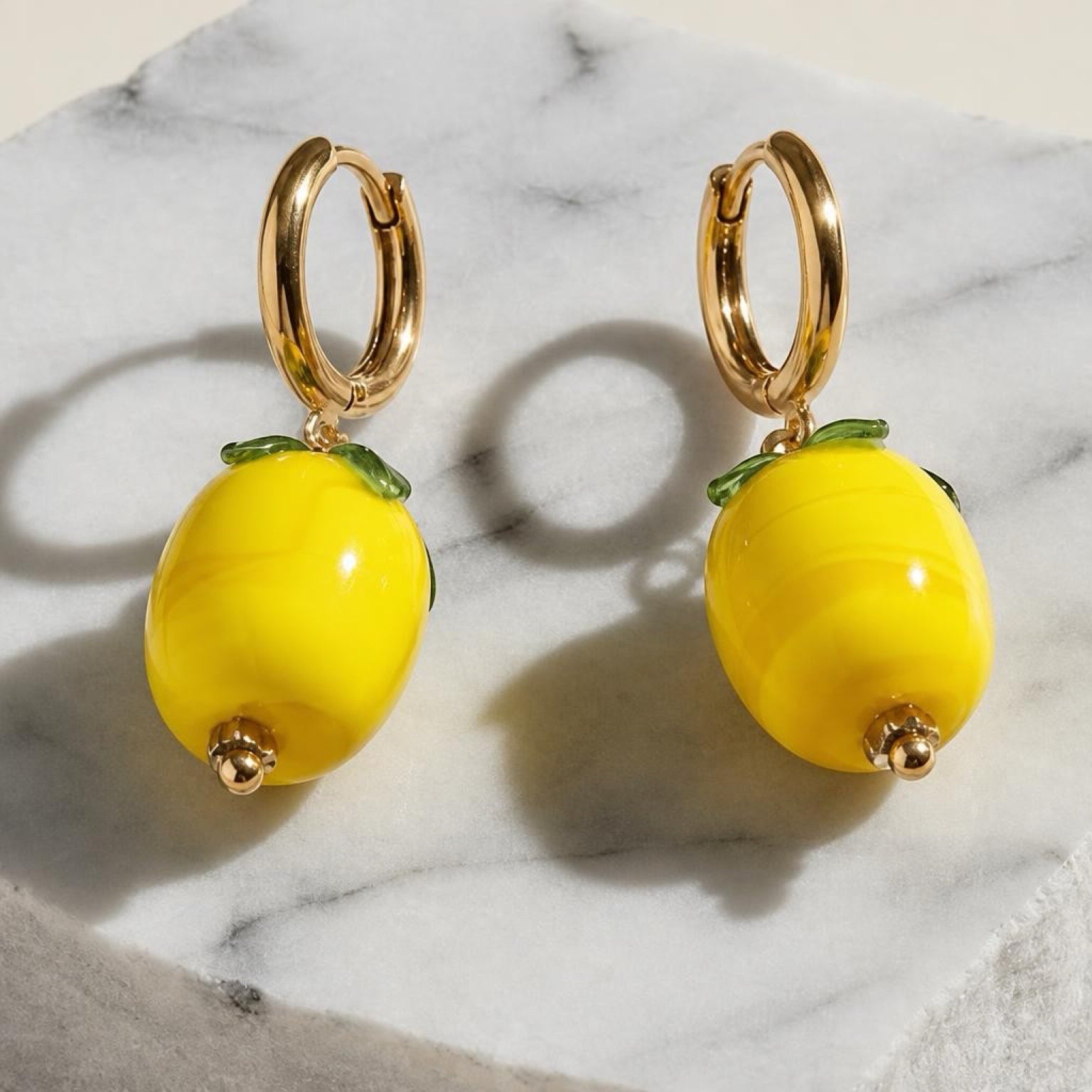 Chévereh gold hoop earrings with glossy yellow glass lemon charms and small green leaves on a marble surface.