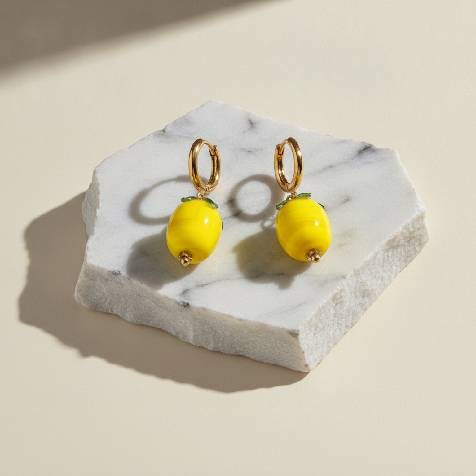 Chévereh gold hoop earrings with glossy yellow glass lemon charms and green leaf details on a marble display