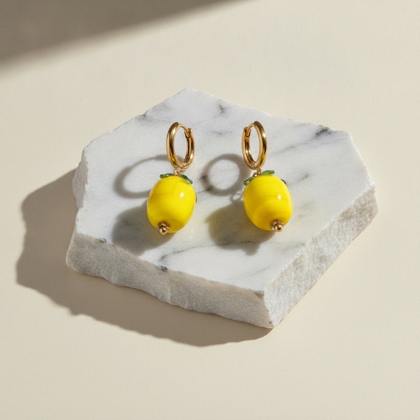 Chévereh gold hoop earrings with glossy yellow glass lemon charms and green leaf details on a marble display