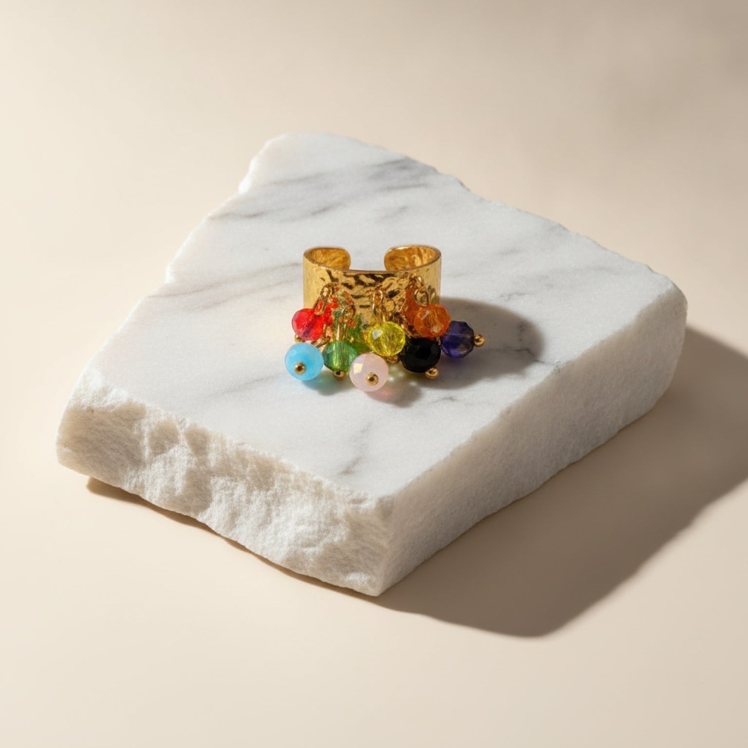 Chévereh gold-toned hammered metal statement ring with dangling multi-colored beaded charms on a marble stone base.