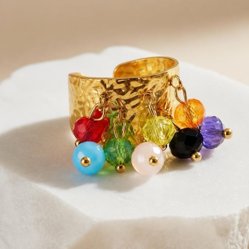 Chévereh gold hammered metal statement ring with dangling multi-colored beaded charms on a white stone surface