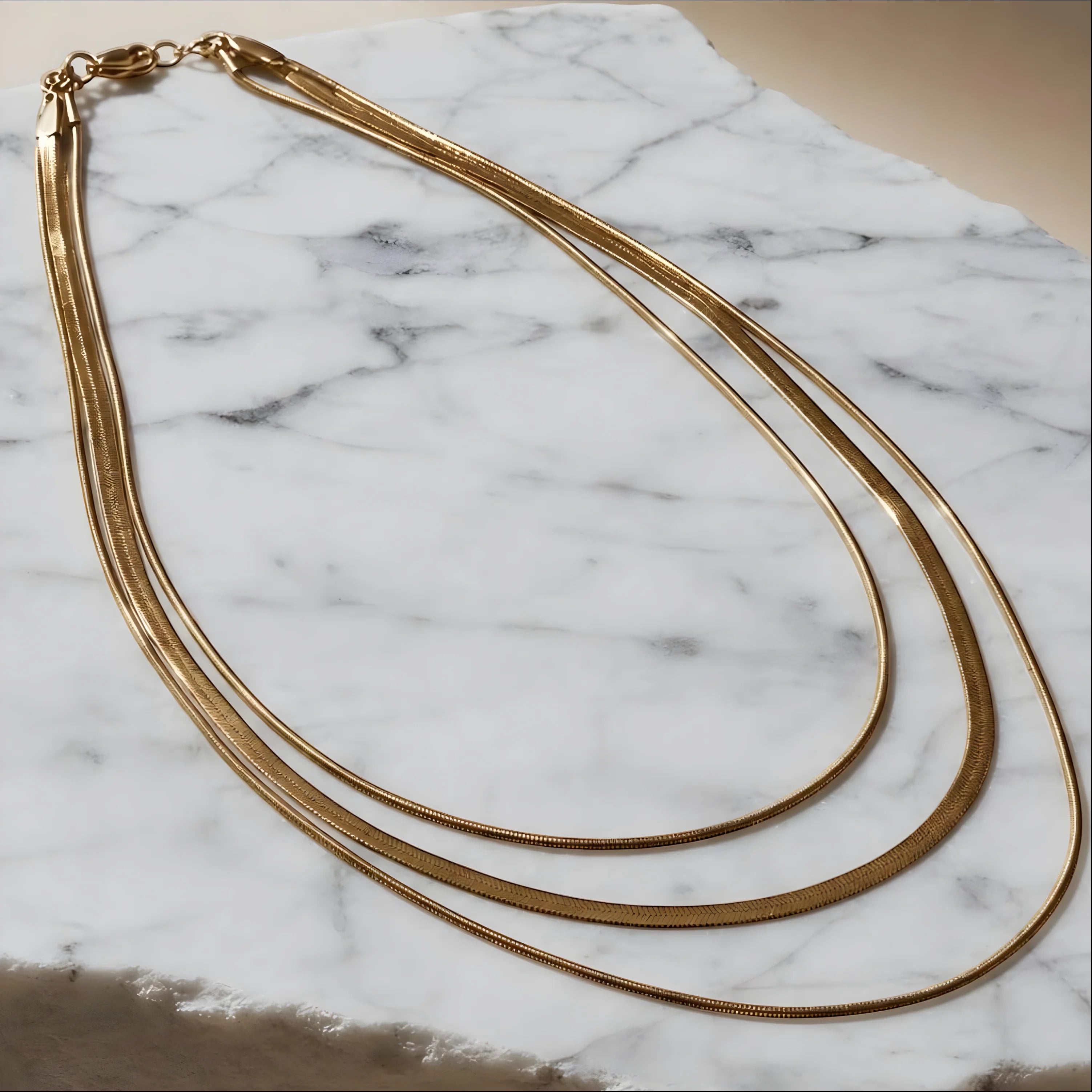 Chévereh gold-tone layered herringbone chain necklace with three delicate strands on a white marble surface.