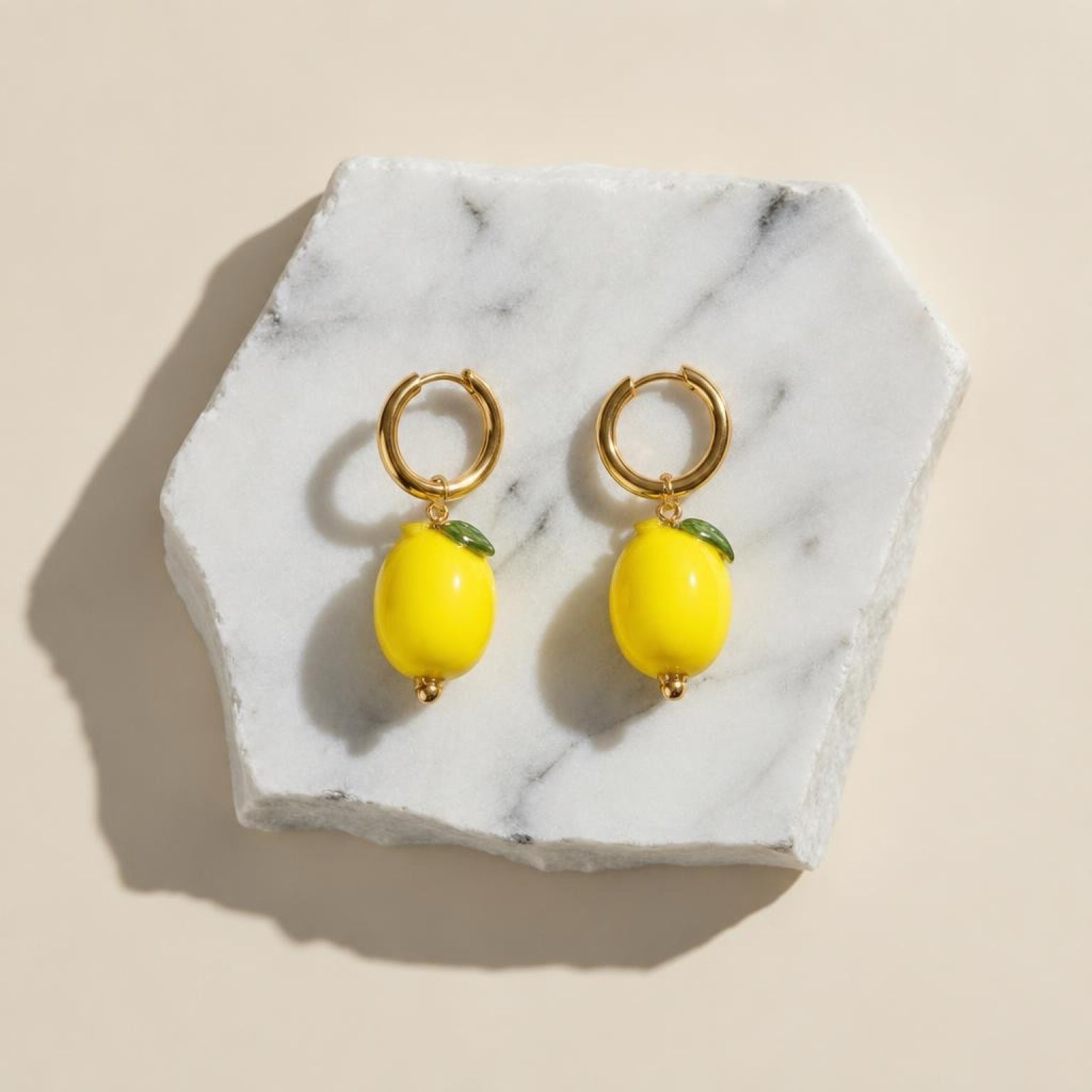 Chévereh gold hoop earrings with glossy yellow lemon charms and green leaf details on a white marble display base.