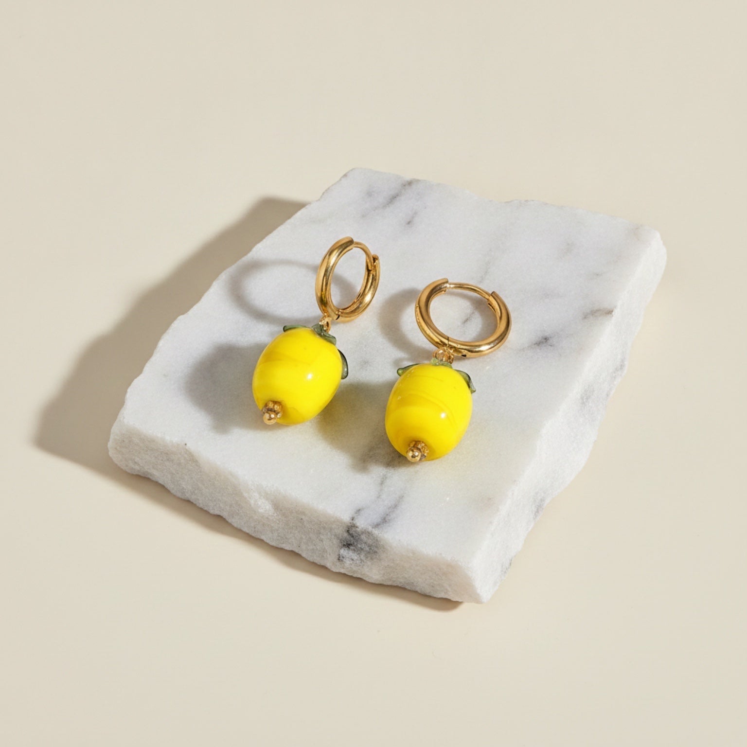 Chévereh gold hoop earrings with glossy yellow lemon charms and green leaf details on a white marble base.