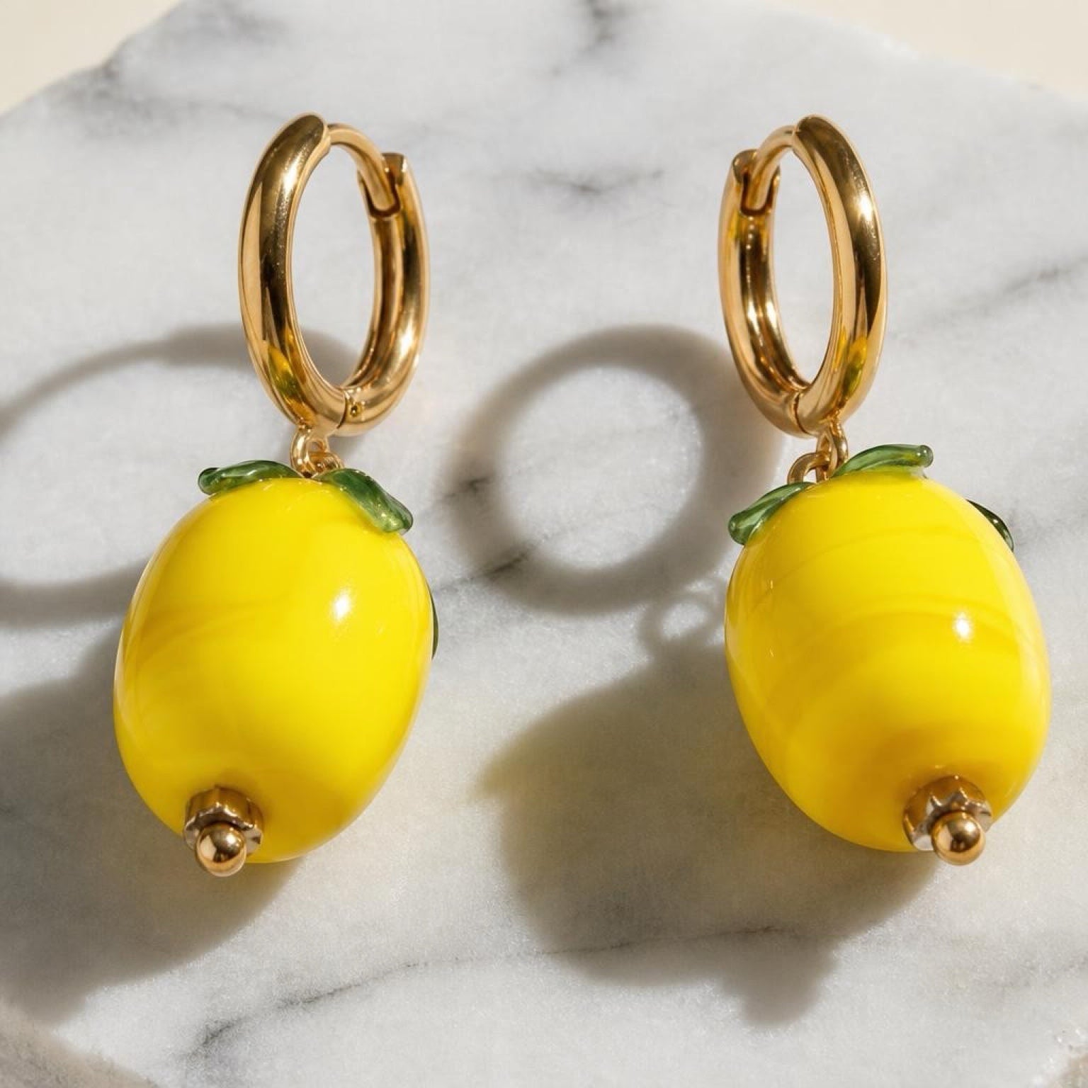 Chévereh gold hoop earrings featuring glossy yellow glass lemon charms with small green leaf accents on marble.