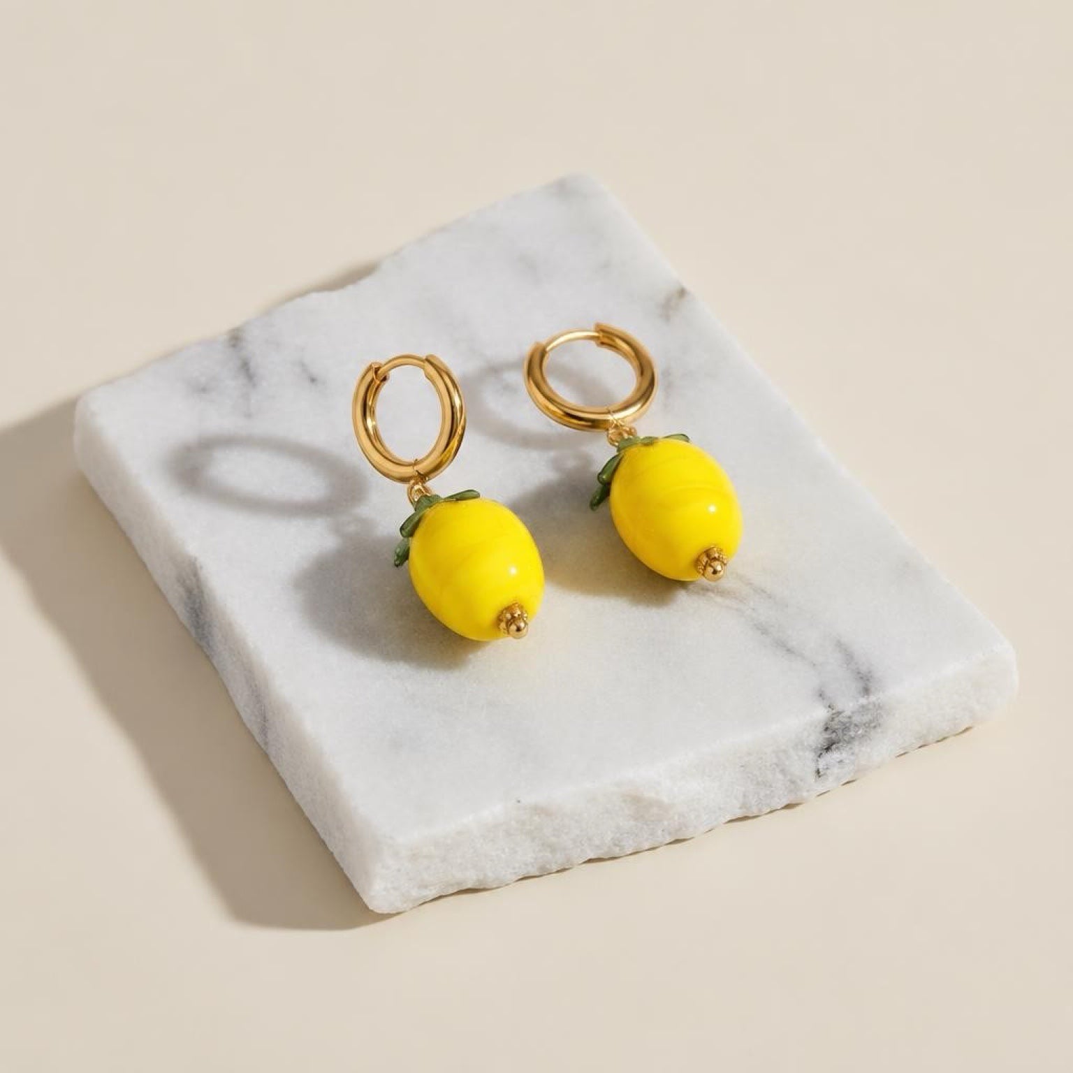 Chévereh gold hoop earrings featuring glossy yellow lemon charms with small green leaf accents on a marble base.