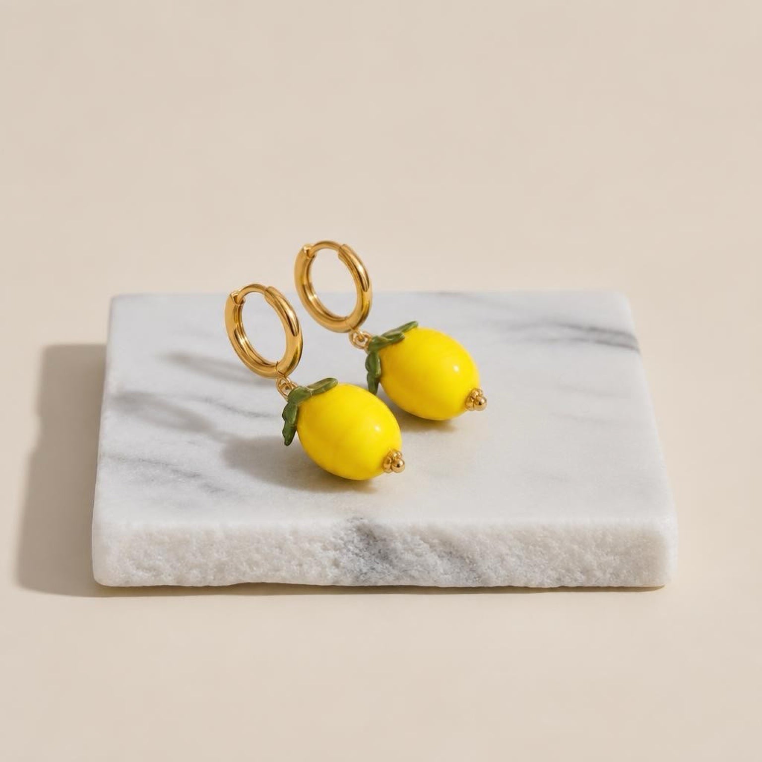 Chévereh gold hoop earrings with glossy yellow lemon charms and green leaf details resting on a marble block